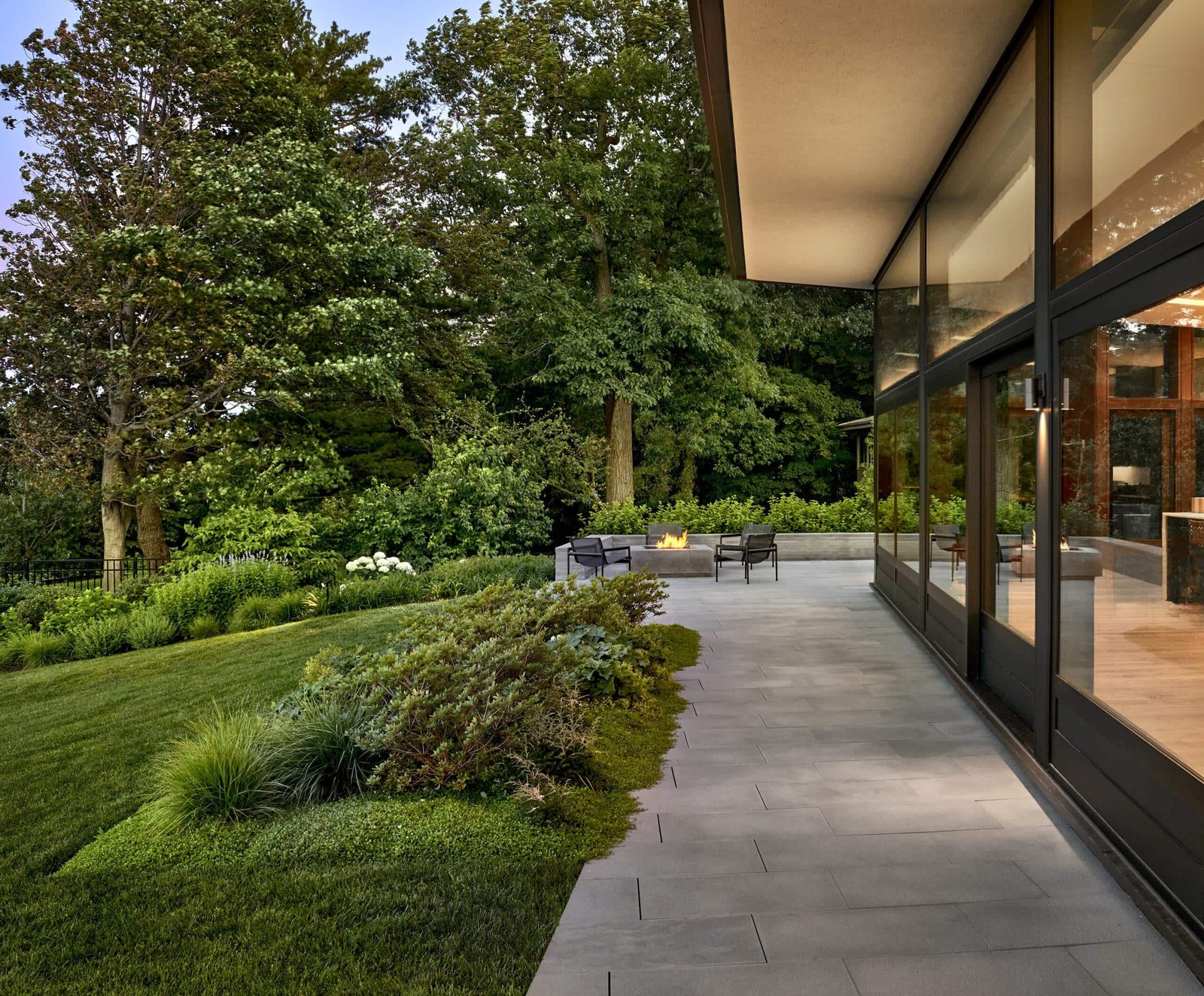 Modern house with large glass windows overlooking a landscaped patio with outdoor seating, surrounded by lush green lawn and trees.