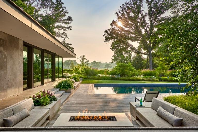 Modern backyard with a rectangular pool, outdoor fire pit, seating area, and a minimalist house with large glass windows overlooking a serene lake.