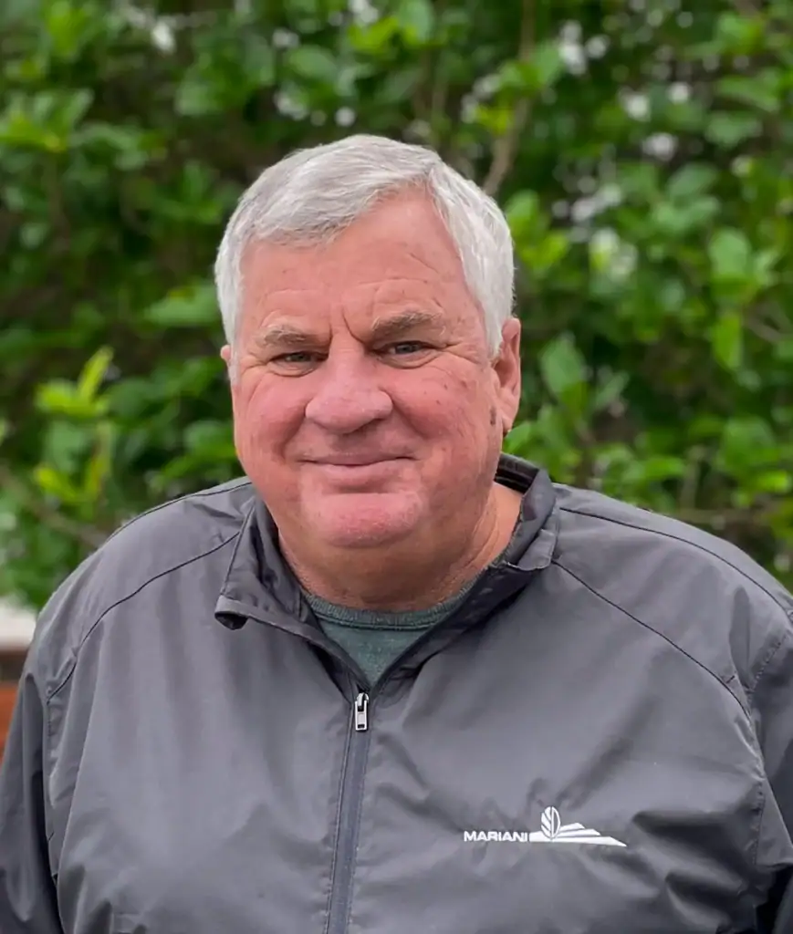 A man with grey hair smiles, wearing a grey jacket with "MARIANI" written on it, standing outdoors with green foliage in the background.