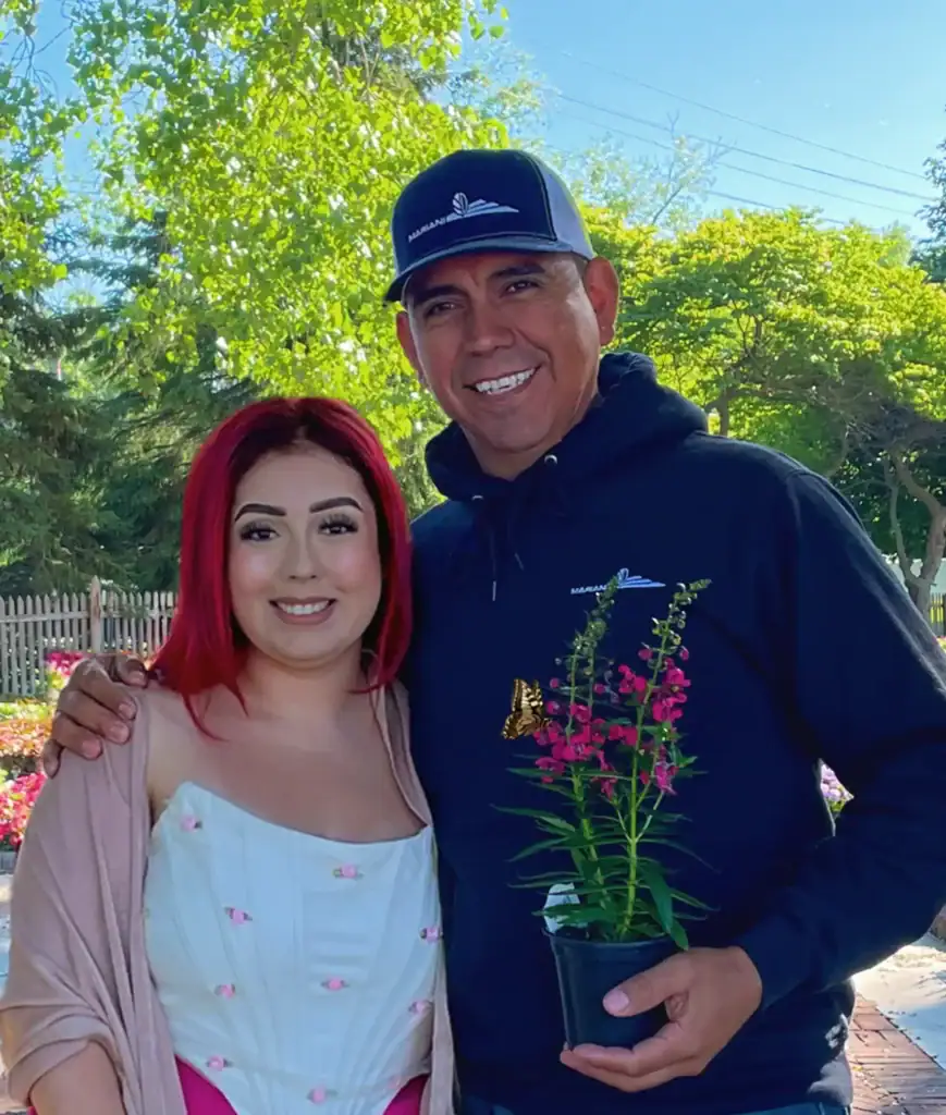 A smiling man and woman stand close together outdoors. The man holds a small potted plant. The woman has red hair and wears a white dress. Trees and a wooden fence are visible in the background.