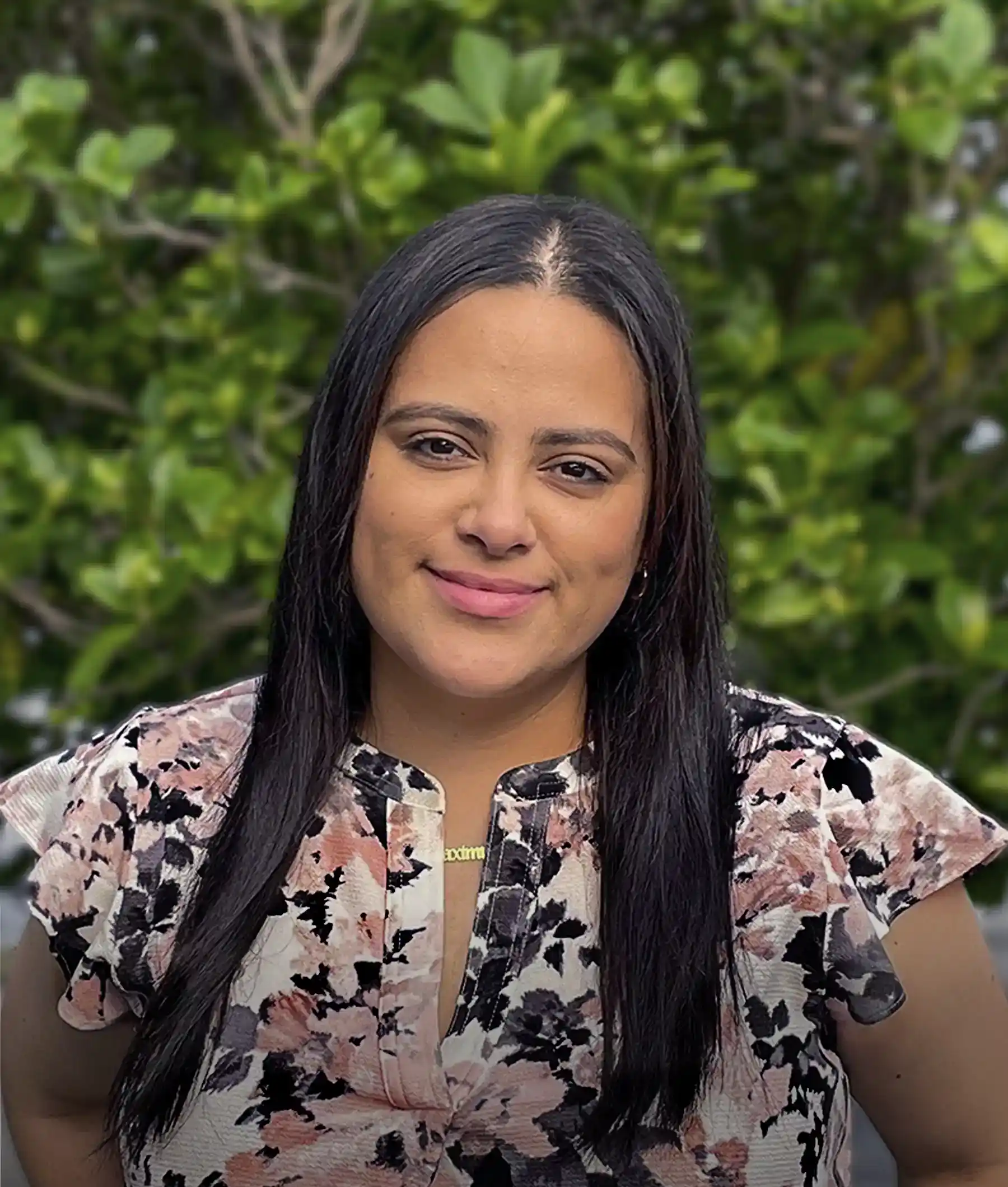 A person with long dark hair and a floral blouse stands in front of a green leafy background, smiling at the camera.