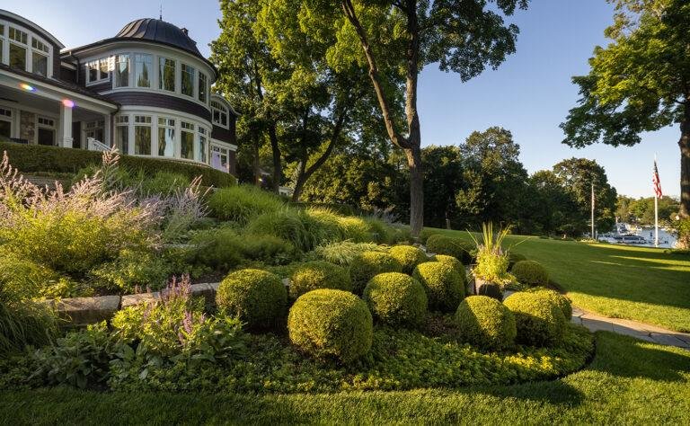 Large house with a rounded turret and numerous windows, surrounded by lush greenery, trimmed bushes, and well-maintained Lake Geneva landscaping. An American flag is visible in the background.