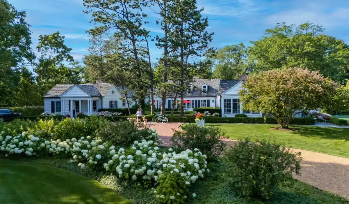 A large, white house with multiple sections and a gray roof is surrounded by trees, flowers, and a manicured lawn under a blue sky. Bicycles and a few people are visible near the entrance.