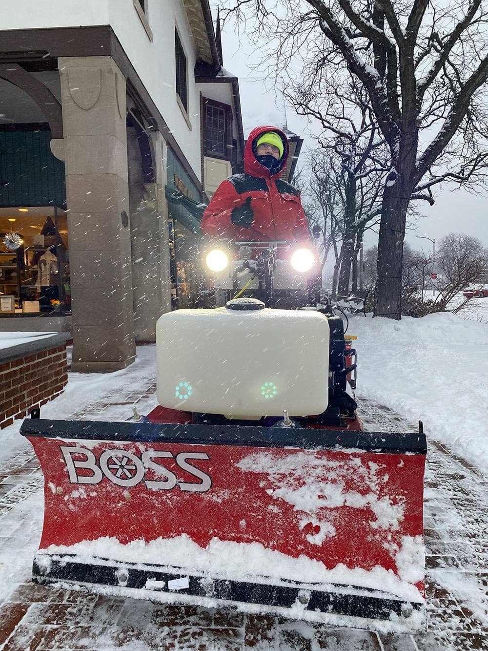 A person in a red jacket expertly manages a red snowplow on a snowy sidewalk near a building, showcasing efficient snow and ice removal for professional campuses as heavy snowfall creates a winter wonderland scene.