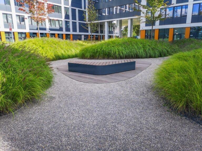 A modern courtyard with a central wooden bench surrounded by well-maintained green grass and gravel paths, adjacent to a building with large windows and orange accents, reflects the meticulous landscape maintenance often required for hospitals.