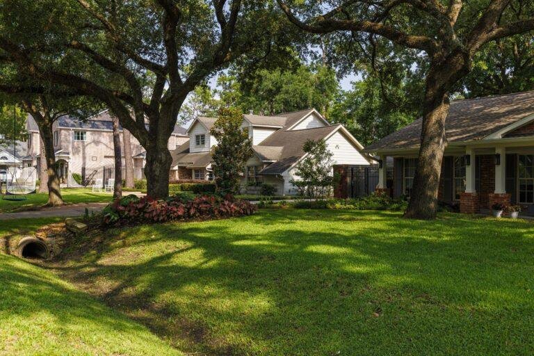 This residential neighborhood boasts green lawns and large trees, perfectly illustrating the benefits of landscape maintenance for HOAs. Several houses line the streets, while a drainage pipe discreetly complements the well-kept grassy area.