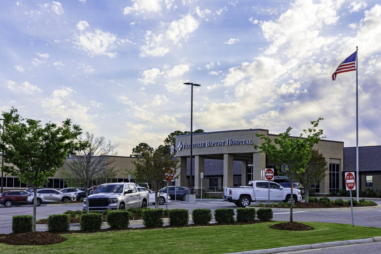 Front view of a hospital building with parked cars and an American flag waving proudly, complemented by well-kept grounds reflecting the meticulous landscape maintenance for hospitals. The sky is partly cloudy, adding a serene backdrop to this inviting scene.