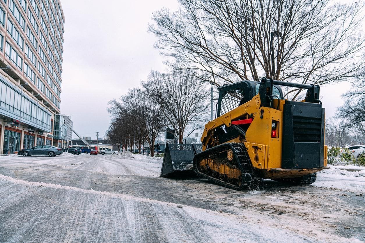 A yellow skid-steer loader, essential for Snow & Ice Removal for HOAs, is parked on a snowy urban street. It's flanked by bare trees and modern buildings in the background.