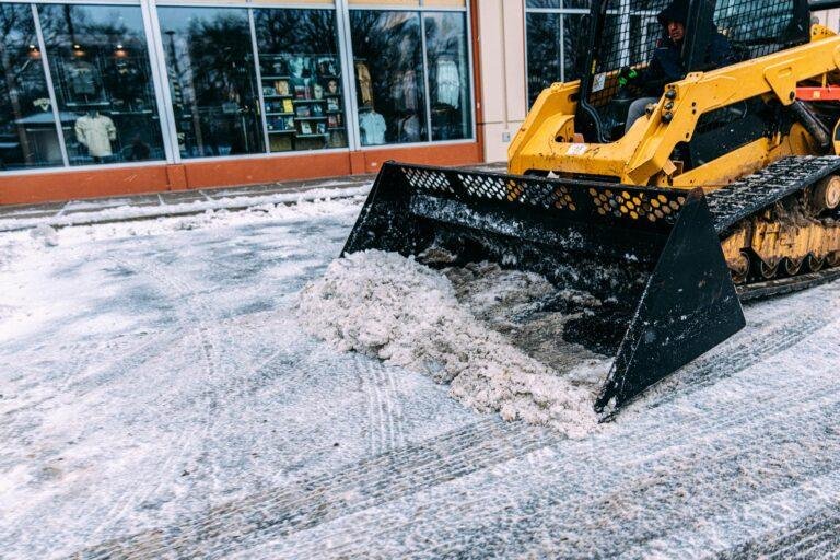 A yellow skid steer loader clears snow from a pavement in front of store windows.