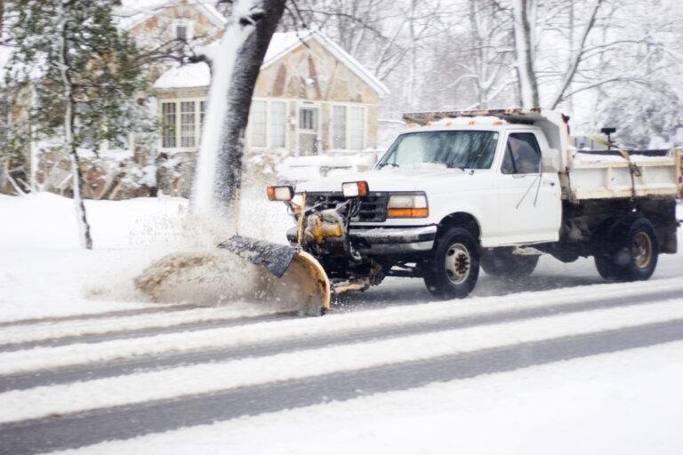 A snow plow truck is expertly clearing snow from a road in a residential area, ensuring safe passage for all, much like the meticulous snow and ice removal required for hospitals. Snow-covered trees and houses stand gracefully in the background.