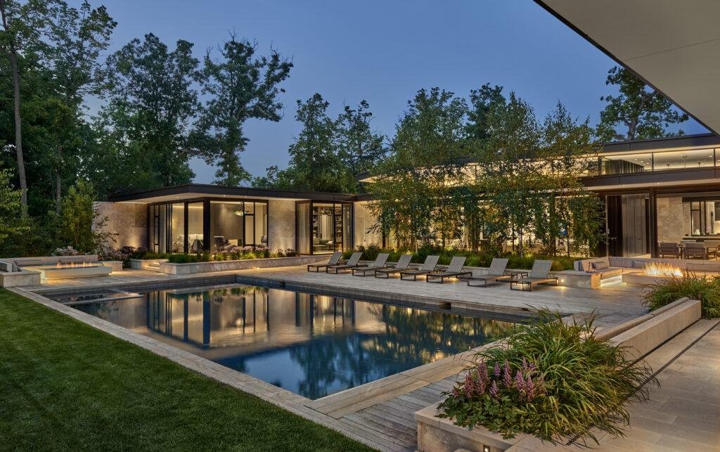 Modern house with large windows and outdoor lighting, featuring a rectangular swimming pool, lounge chairs, and landscaped greenery at dusk.