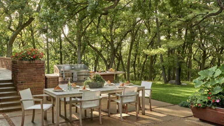 Outdoor dining area with a set table, six white chairs, a grill, and lush greenery—perfect for enjoying expertly designed landscaping in Barrington IL. Steps and a brick wall add charm on the left.