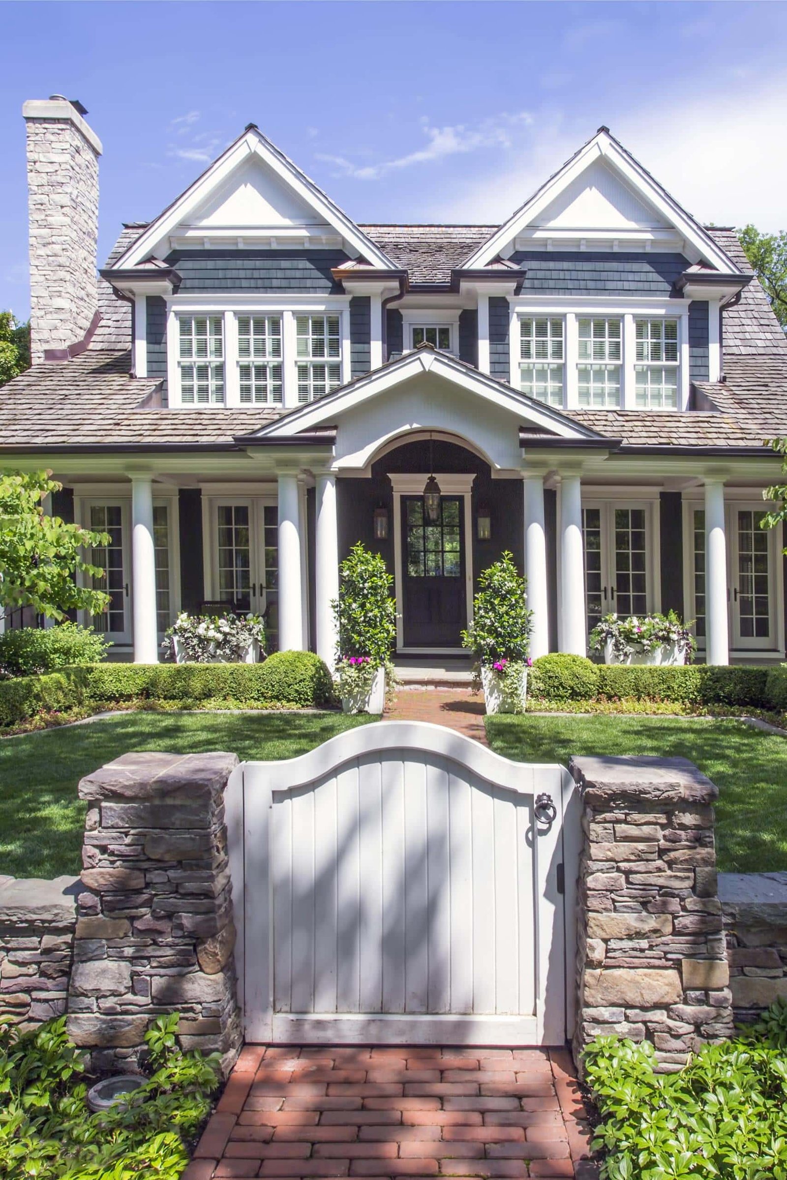 Two-story house with white columns, dark blue siding, and peaked roof, seen behind a stone wall and white gate, features expert landscaping in Hinsdale, IL for a beautifully manicured front yard.