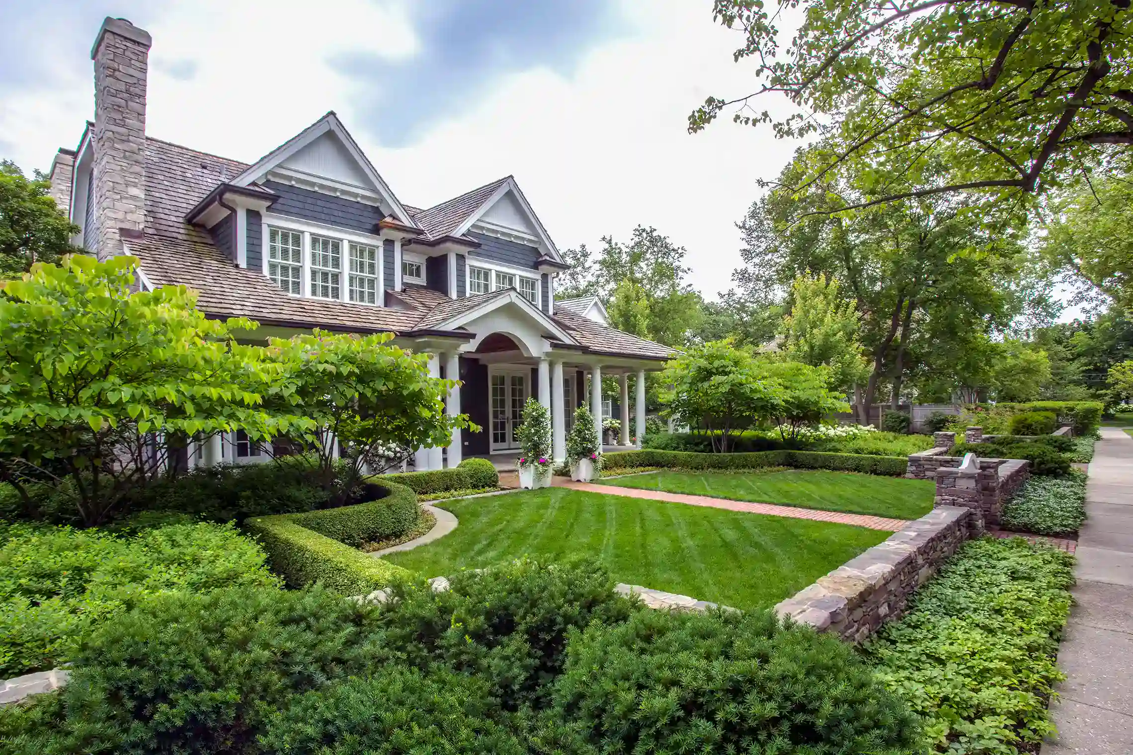 A two-story house with dark siding and white trim is surrounded by manicured hedges, green lawn, and mature trees along a stone-lined sidewalk.