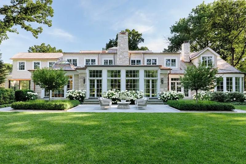 A large, two-story house with many windows, surrounded by green lawn, trees, and neatly landscaped flowerbeds with white flowers. Four chairs and a table are arranged on a stone patio.