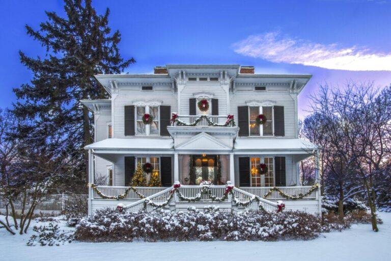 A large, two-story white house decorated with holiday wreaths and garlands, surrounded by snow-covered bushes and trees at dusk.