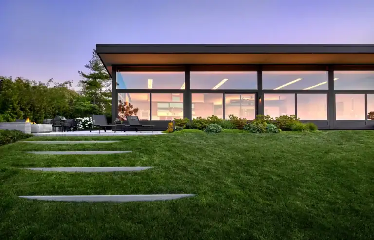 Modern house with large glass windows, a neatly manicured lawn, stone steps, and an outdoor seating area at sunset.