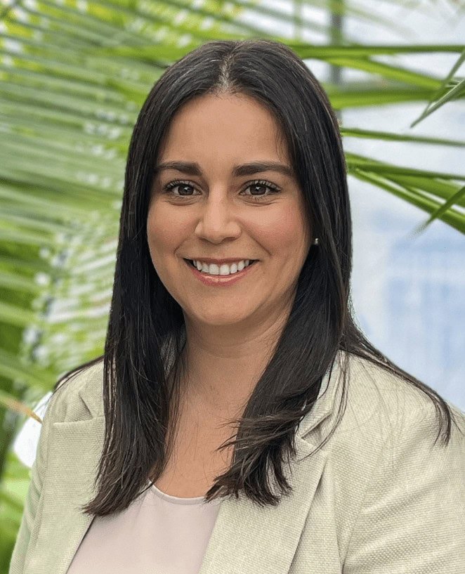 A woman with long dark hair and a light blazer smiles at the camera, standing in front of green palm leaves and a blurred background.