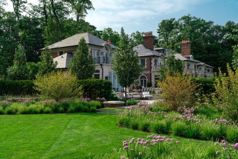 A large brick house with tall chimneys sits behind landscaped gardens with green grass, bushes, and blooming purple flowers on a sunny day.