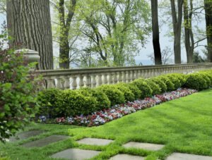 Neatly trimmed hedges and colorful flowers line a stone balustrade on a well-manicured lawn, with tall trees and a distant view in the background.