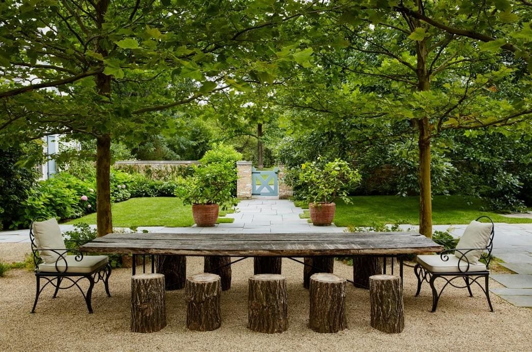 A rustic outdoor dining area features a long wooden table, log stools, cushioned chairs, and is shaded by two leafy trees in a lush garden setting.