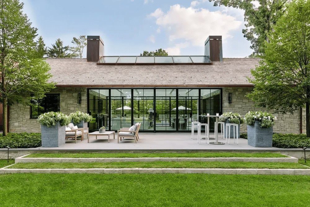 Contemporary stone home with large glass doors opening to a patio furnished with lounge seating, tall planters, and a neatly edged lawn.