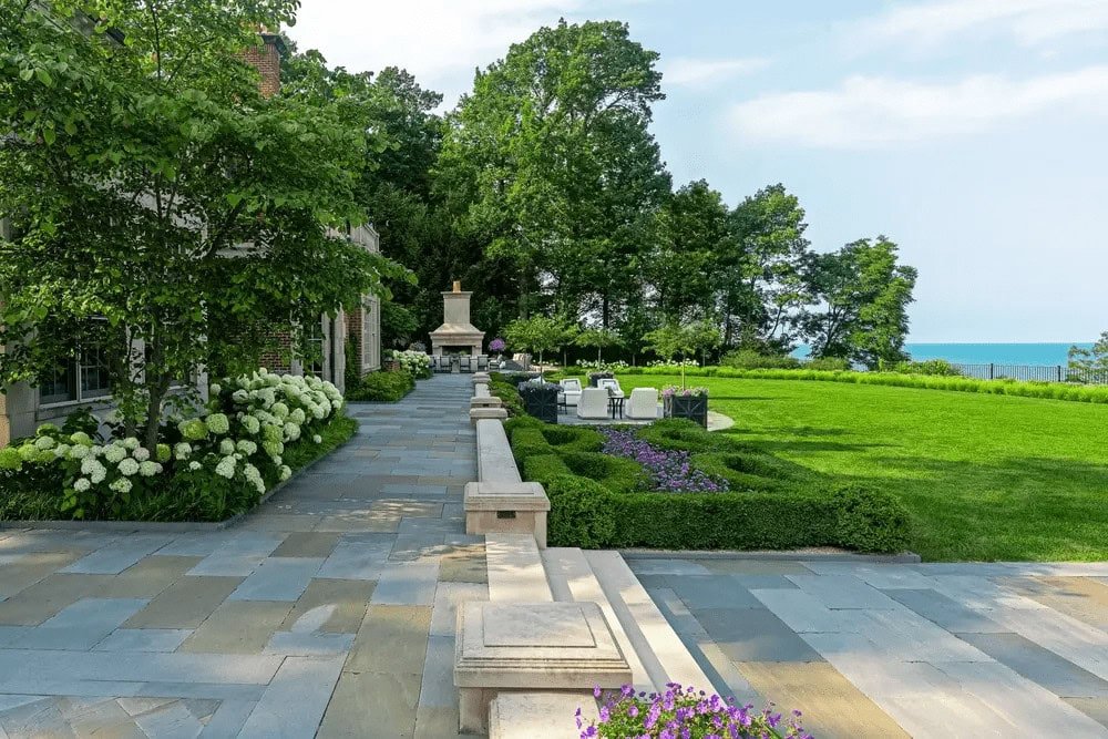 Expansive stone terrace with outdoor seating and fireplace facing a wide green lawn and lake bordered by mature trees.
